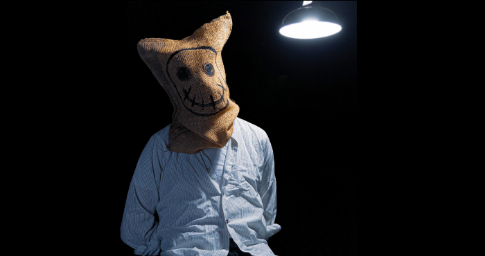 A man sits tied to a chair, under an interrogation lamp, with a sack over his face. The sack has a creepy smiley face drawn on it.